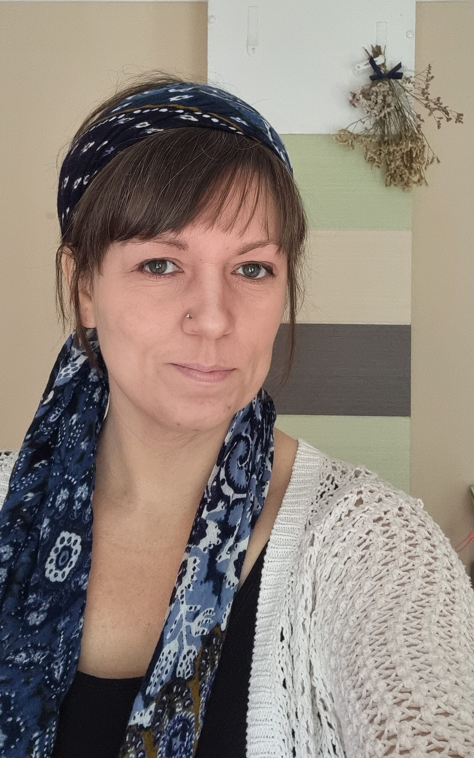 A white woman with brown hair, tied back under a long blue headscarf, and a fringe. I have a nosering and am looking at the camera with a slight smile - more with the eyes than the teeth.